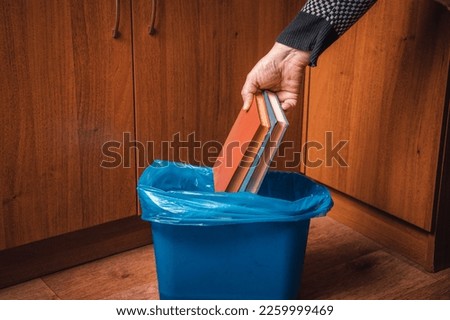 A man throws paper books into a trash can. Man's hand with old books and trash can in blue Royalty-Free Stock Photo #2259999469