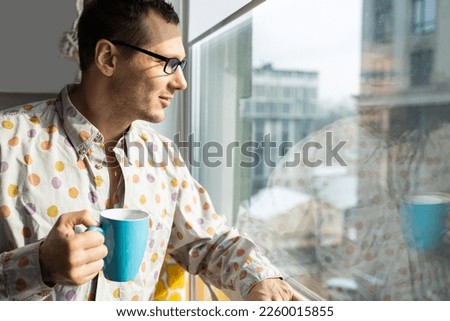 Smiling young adult businessman holding drinking coffee cup in office entrepreneur looking through window while standing in modern workplace man taking a time off or taking a brake from hard work Royalty-Free Stock Photo #2260015855