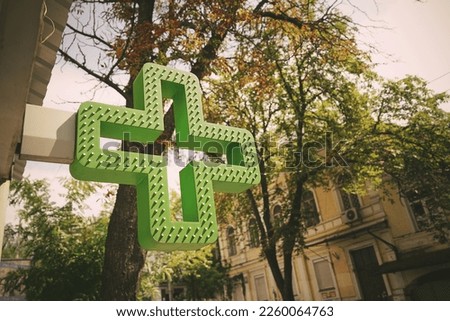 Green medical cross sign fixed on pharmacy facade outside. Drug store signage hanging on building wall at city street.