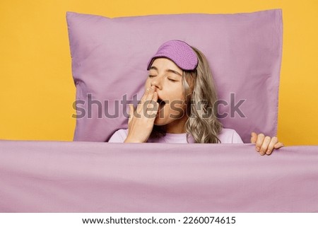 Young woman wears purple pyjamas jam sleep eye mask rest relax at home lies wrap under blanket duvet yawn cover mouth isolated on plain yellow background studio portrait. Good mood night nap concept
