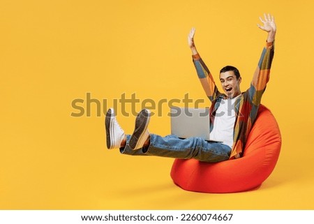 Full body young middle eastern man he wear casual shirt white t-shirt sit in bag chair IT woman hold use work on laptop pc computer raise up hands finish job isolated on plain yellow background studio