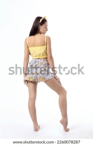 A young woman with dark hair wearing a beach suit stands with her back against a white background.