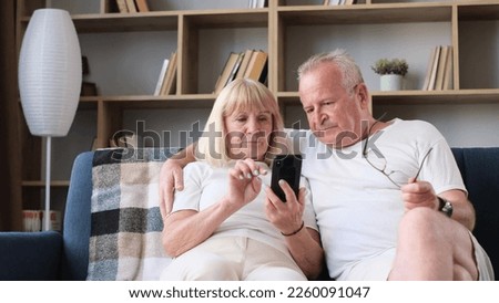 Modern retired couple sitting on sofa and using mobile apps. Grandfather and grandmother with a smartphone in their hands. Modern mobile phones Royalty-Free Stock Photo #2260091047