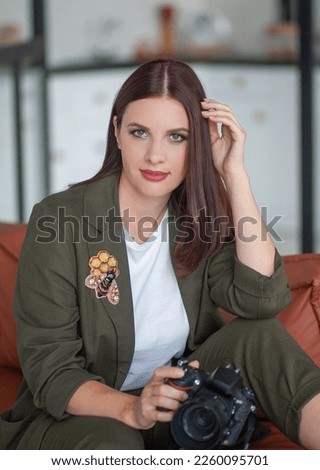 Beautiful young woman is sitting on the sofa at home. A camera is in the hands of a woman. Selfie photography. Portrait of a lovely and beautiful professional photographer.
