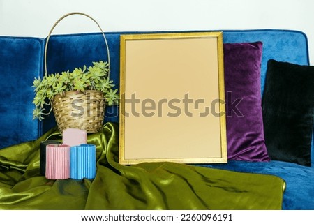 A golden frame, some plants, some candles, fabrics and cushions on a velvet sofa