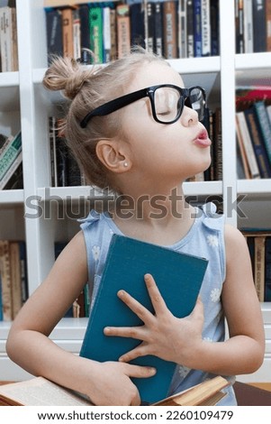 Little cute girl in the glasses sitting in the front of bookshelf. Concept of education, soft focus background
