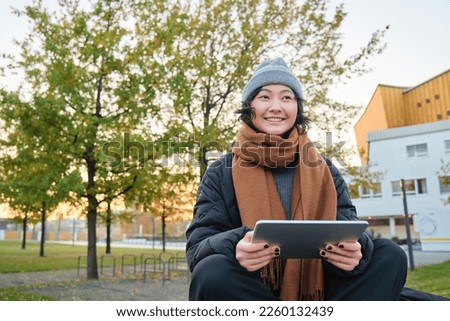 Portrait of asian girl in warm clothes, sits on bench with digital tablet and graphic pen, smiling happily, draws outdoors in chilly weather.