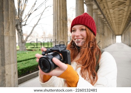 Young redhead female photographer, makes lifestyle shooting in city centre, takes photos and smiles, looks for perfect shot, makes picture.