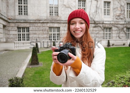 Redhead girl photographer takes photos on professional camera outdoors, captures streetstyle shots, looks excited while taking pictures.