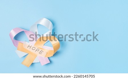 Three ribbons pink, white and yellow with sticky tape with the word hope lie on the left on a blue background with copy space on the right, flat lay close-up. World cancer day concept.