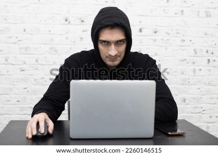 Young confident guy in hoodie sitting at the table and working on laptop