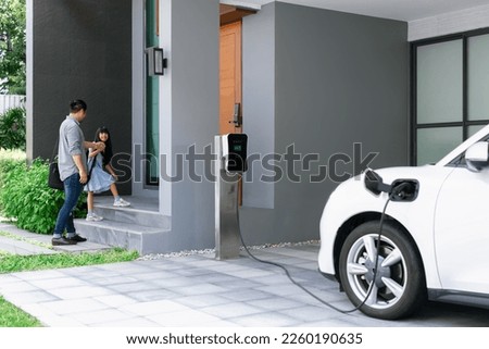 Progressive father and daughter plugs EV charger from home charging station to electric vehicle. Future eco-friendly car with EV cars powered by renewable source of clean energy. Royalty-Free Stock Photo #2260190635