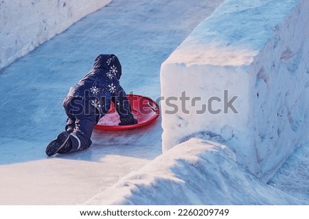 In winter on an ice slide. A boy in a hooded jacket holding a red plastic plate in his hand. Side view. Bright sunlight. Ice boat for winter fun.