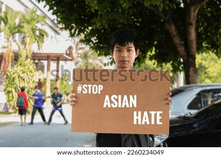 Asian boy holds brown protesting paper which has texts "#StopAsianHate", blurred school area background, concept for calling people to stop bullying, violence and hating Asian people all the world.