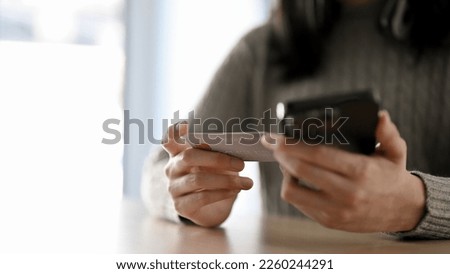Cropped image of an Asian woman in cozy sweater holding a credit card and a smartphone at the desk. mobile banking, online payment, online money transfer