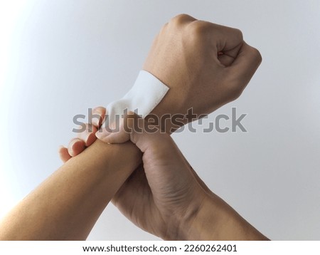 A person with a sprained wrist, with an anti-inflammatory, pain relief  patch on the left wrist,  and the other hand hold on it, in front of an all-white background.