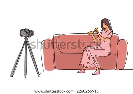 Single continuous line drawing stylish beauty blogger is sitting at sofa, reviewing heels shoe in her hands while recording video with digital camera, tripod. One line draw design vector illustration