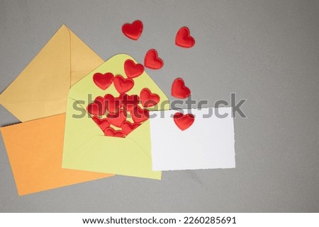 Light green, orange, yellow envelopes and a white card for text with red falling hearts on a gray concrete background. Happy Valentine's Day greeting, background. Flat lay