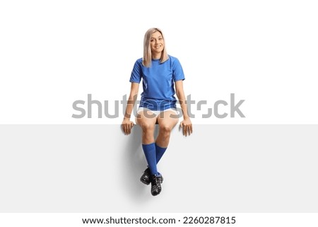 Full length portrait of a female football player sitting on a blank white panel isolated on white background