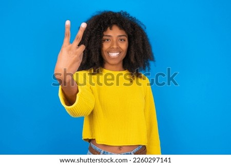 young woman with afro hairstyle wearing yellow sweater against blue background smiling and looking friendly, showing number two or second with hand forward, counting down Royalty-Free Stock Photo #2260297141