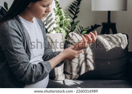 Selective focus of sad pregnant woman suffering pain on hands and fingers, arthritis inflammation. Upset caucasian female has pain in wrist sitting on couch at home. Health care and medical concept