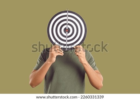 Successful male freelancer who in search of target audience covers his face with darts board. Unknown young man in casual T-shirt hides his face behind darts on khaki background. Targeting concept. Royalty-Free Stock Photo #2260331339