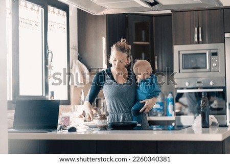 Happy mother and little infant baby boy together making pancakes for breakfast in domestic kitchen. Family, lifestyle, domestic life, food, healthy eating and people concept Royalty-Free Stock Photo #2260340381