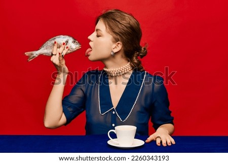 Tasting new taste. Young extravagant woman with cup of coffee and raw fish on bright red background. Delicious weird taste. Retro style. Complementary colors. Food pop art photography. Royalty-Free Stock Photo #2260343193
