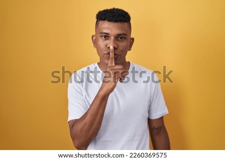 Young hispanic man standing over yellow background asking to be quiet with finger on lips. silence and secret concept. 