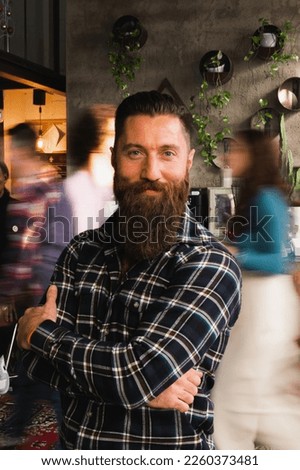 Vertical portrait of smiling man with beard and crossed arms standing looking at camera. Unrecognizable blurred people walking.