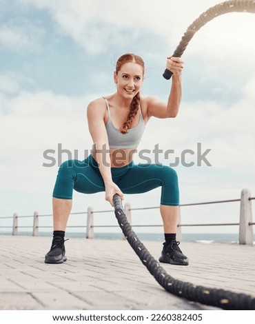 Woman, battle rope exercise and smile at ocean park for fitness, strong body and wellness at outdoor training. Girl, focus and workout by sea with goal, health or motivation for muscle development Royalty-Free Stock Photo #2260382405
