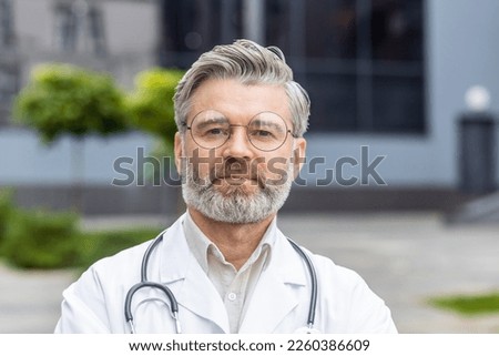 Close-up portrait of mature man in medical coat, senior doctor thinking and looking at camera near clinic outside. Royalty-Free Stock Photo #2260386609