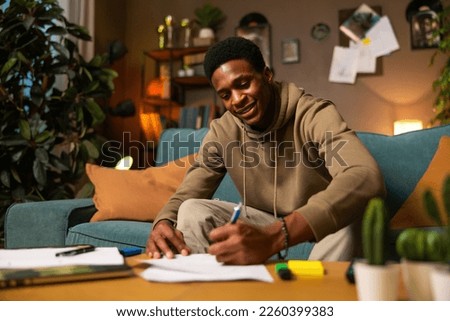 Attractive american teenage student wearing casual new clothes sitting in the living room writing studying preparing for exams at the university making notes in copybook pad. Royalty-Free Stock Photo #2260399383