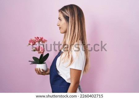 Young blonde woman wearing gardener apron holding plant looking to side, relax profile pose with natural face and confident smile. 
