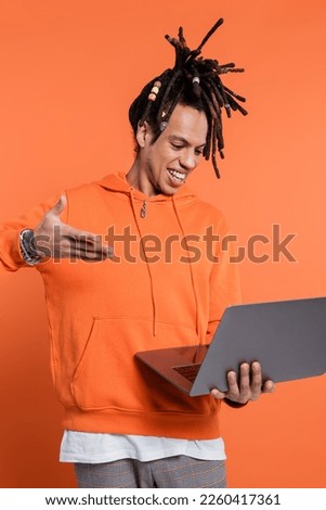 happy multiracial man with dreadlocks and tattoo pointing at laptop on coral background 