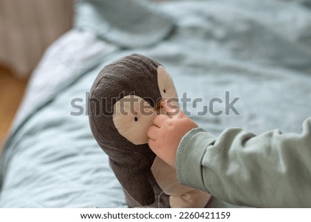 Baby's hand shows and touches the beak of a toy penguin, background with space for text.

