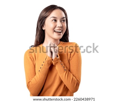 A studio portrait of a young Asian Indonesian woman wearing an orange long-sleeve shirt looks happy as she smiles while her hands are clasped in front of her chest. Isolated on a white background. Royalty-Free Stock Photo #2260438971