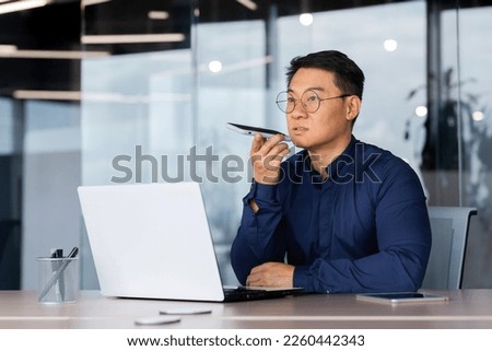 Successful asian man inside office working with laptop, businessman recording audio message using app on smartphone, man using artificial intelligence assistant to help finding a solution. Royalty-Free Stock Photo #2260442343
