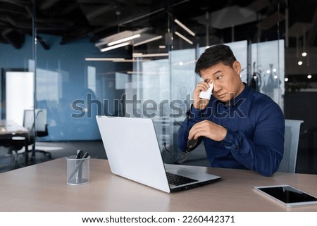 Watery eyes of man inside office, businessman overtired and overloaded with work hurts eyes, Asian man at work with laptop in glasses and shirt near window. Royalty-Free Stock Photo #2260442371