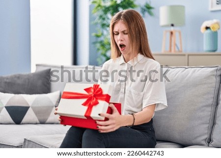 Beautiful woman holding gift in shock face, looking skeptical and sarcastic, surprised with open mouth 