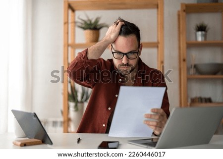 Overworked businessman at home office looking at paper document holding hand on his head feeling hopeless trying to find solution for given problem working too hard need break for better concentration Royalty-Free Stock Photo #2260446017