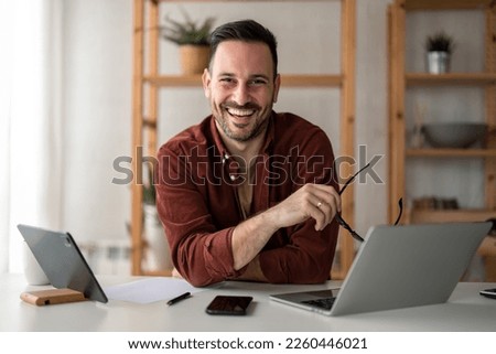 Smiling confident businessman looking at camera sitting at home office desk. Modern stylish corporate leader, successful manager or small business owner holding glasses posing for business portrait. Royalty-Free Stock Photo #2260446021