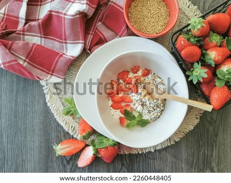 Breakfast view, with a bowl of natural yogurt with pieces of strawberry, mint, linseed and chia seeds; background with whole strawberries, bowl with linseed and kitchen towels on dark wooden table... Royalty-Free Stock Photo #2260448405