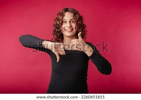 Cheerful redhead woman wearing black ribbed dress isolated over red background making thumbs up and thumbs down hands sign. Looking at the camera.