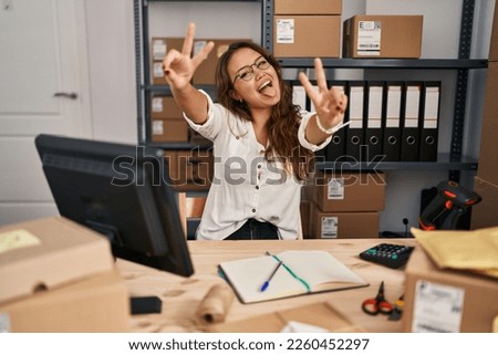 Young hispanic woman working at small business ecommerce smiling with tongue out showing fingers of both hands doing victory sign. number two. 