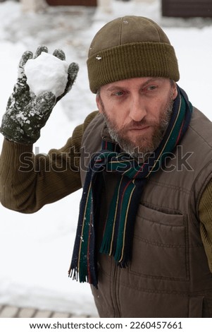 Snow games in the air. A man with a beard in a warm vest and a hat throws snowballs in the yard of a private house on a frosty day

