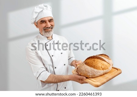 Chef-cooker in chef's hat and jacket working in bakery, holding French bread board with bread. Senior professional baker man wearing chef's outfit. Character kitchener, pastry chef for advertising. Royalty-Free Stock Photo #2260461393