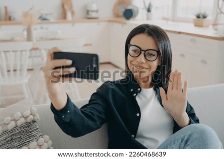 Smiling young female greeting friend, waving hand, having video conversation by smartphone sitting on couch at home. Businesswoman wearing glasses chatting by videocall make hello gesture, welcoming.