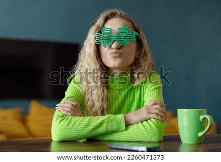 A young woman in green clover-shaped glasses looks in surprise at a laptop screen at a table in the room. Holds a cup with a drink for St. Patrick's Day