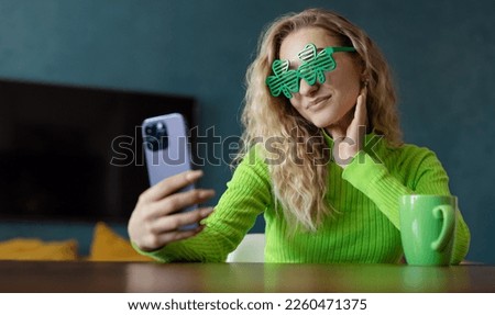 A young woman in green clover-shaped glasses looks in surprise at a smartphone screen at a table in the room. Holds a cup with a drink for St. Patrick's Day